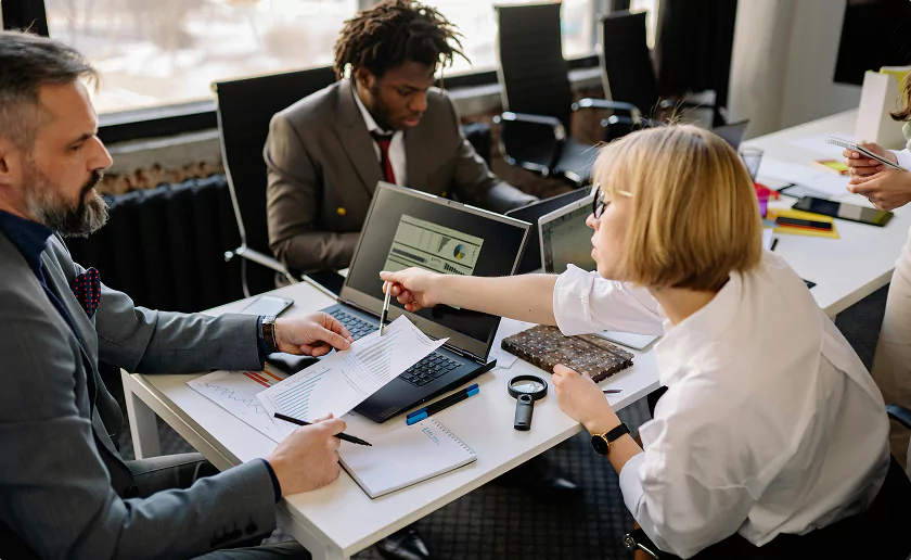 Diverse team of professionals in a modern office analyzing financial reports and data on laptops, collaborating closely at a meeting table.