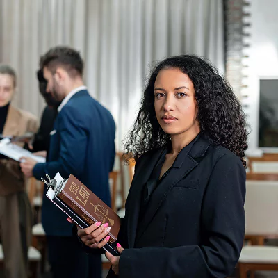 Professional woman in a black blazer holding legal documents, standing confidently in a courtroom or formal meeting space with colleagues in the background.