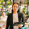 Young businesswoman holding a tablet, standing in a vibrant office with plants and coworkers in the background.