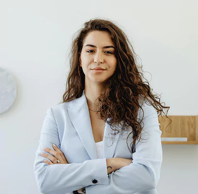 Confident young woman with curly hair wearing a light blue blazer, standing with arms crossed in a bright, minimalistic office
