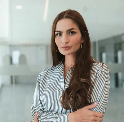 Professional woman with long brown hair wearing a striped shirt, standing confidently with arms crossed in an office setting.