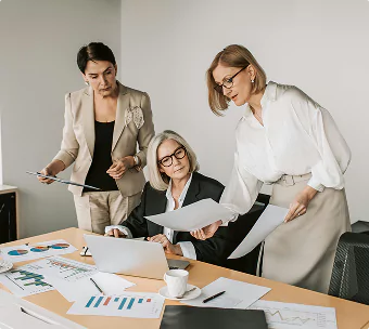 Three women are gathered around a table, discussing documents and data in a professional office setting.