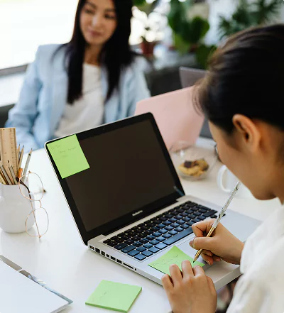A woman writes on a sticky note at her laptop, while another person sits across the table.