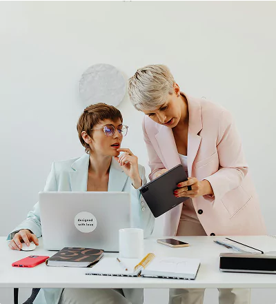 Two women in pastel suits collaborate at a desk, reviewing content on a tablet and laptop.